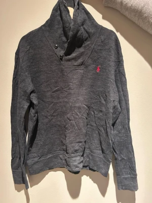 Mörkgrå hoodie från Polo Ralph Lauren - Mörkgrå hoodie från Polo Ralph Lauren med hög krage och knappdetaljer. Klassisk röd logga broderad på bröstet. Tillverkad i mjukt material, perfekt för en avslappnad stil. Långa ärmar och ribbade muddar för extra komfort.