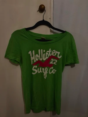 Grön Hollister t-shirt med tryck - Snygg grön t-shirt från Hollister med stort vitt och rosa tryck på bröstet där det står 'Hollister Surf Co 22'. Klassisk rund halsringning och korta ärmar💗