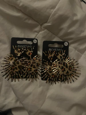 Guldiga statementörhängen Montini - Säljer två par stora statementörhängen från Montini i guld med leopardmönster. Örhängena har en unik solfjäderform med spretiga detaljer som verkligen drar blickarna till sig. Perfekta för dig som vill sticka ut och addera lite edge till din stil. Säljer en för 100kr och 2 för 200kr