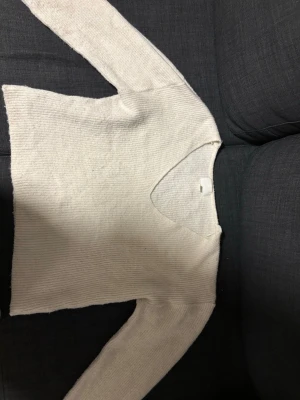 Vit stickad tröja med v-ringning - Mysig vit stickad tröja med v-ringad hals och långa ärmar. Tröjan har en enkel, rak passform och är tillverkad i ett mjukt, ribbat material som känns skönt mot huden. Perfekt att matcha med jeans eller kjol för en avslappnad look.