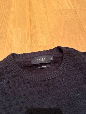 Mörkblå stickad tröja från Riley - Snygg mörkblå stickad tröja från Riley med diskret randig struktur. Tröjan har rund halsringning och långa ärmar. Tillverkad i en skön blandning med catskill linen för extra komfort och stil.