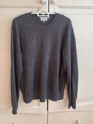 Mörkgrå stickad tröja från Sandö - En stilren mörkgrå stickad tröja från Sandö med rund halsringning och långa ärmar. Tröjan har ribbade muddar vid ärmslut och nederkant, vilket ger en klassisk och clean look. Perfekt att matcha med jeans eller chinos.