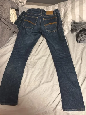 Nudie Mörkblå jeans med gul söm - Snygga mörkblå jeans från nudie med klassiska raka ben och coola guldfärgade sömmar på bakfickorna. Jeansen har fem fickor och en läderpatch vid linningen. Perfekt för dig som gillar stilrena och tidlösa jeans.