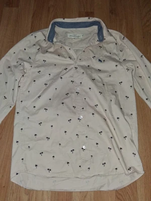 Beige skjorta med palmmönster - Snygg beige skjorta med små svarta palmer broderade över hela tyget. Skjortan har klassisk krage med kontrasttyg i blått, knappar framtill och långa ärmar. Perfekt för dig som vill ha en chill men ändå stilren look.