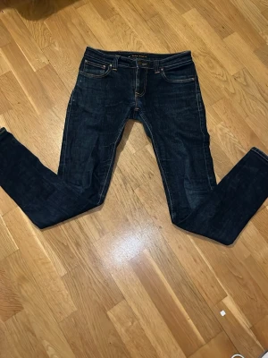Mörkblå jeans från Nudie Jeans - Snygga mörkblå jeans från Nudie Jeans med klassisk femficksdesign och raka ben. Jeansen har kontrastsömmar och detaljer i kopparfärgad metall. Perfekta för en avslappnad och stilren look.