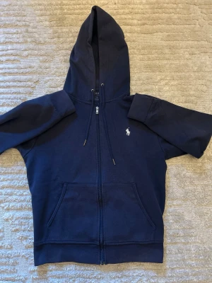 Mörkblå hoodie från Polo Ralph Lauren - Mörkblå hoodie med dragkedja från Polo Ralph Lauren. Klassisk design med huva och dragsnören, två stora fickor framtill och den ikoniska vita logon broderad på bröstet. Tillverkad i mjuk bomullsblandning för maximal komfort.