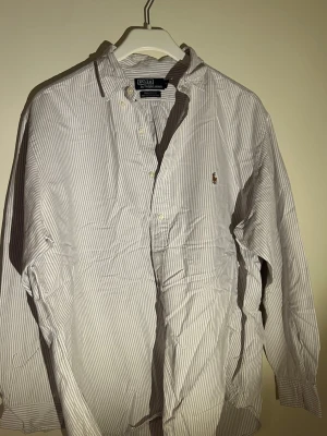  Regnbåg randig skjorta från Polo Ralph Lauren - Klassisk långärmad skjorta från Polo Ralph Lauren i vitt med tunna grå ränder. Skjortan har button-down krage, broderad logga på bröstet och är tillverkad. Perfekt för en clean och stilren look.