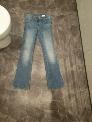 Blå bootcut jeans med slitningar - Säljer ett par blå bootcut jeans med snygga slitningar framtill och klassiska fem fickor. Jeansen har en ljus tvätt och utsvängda ben, perfekta för en avslappnad och trendig look. Materialet är denim i bomull.