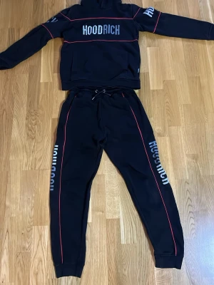 Svart HOODRICH hoodie och byxor set - Säljer ett svart loungewear set från HOODRICH med matchande hoodie och byxor. Setet har röda detaljer längs ärmar och ben samt HOODRICH-logga i vitt på bröstet, ärmen och båda benen. Hoodien har huva och byxorna har dragsko i midjan. Pris kan diskuteras 