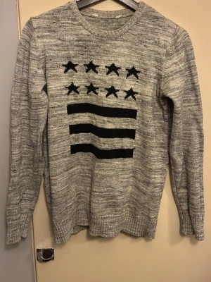 Grå stickad tröja med stjärnor - Gråmelerad stickad tröja med svart mönster av stjärnor och ränder på bröstet. Tröjan har rund halsringning, långa ärmar och ribbstickade muddar vid ärmslut och nederkant. Perfekt för dig som gillar en avslappnad och trendig stil.