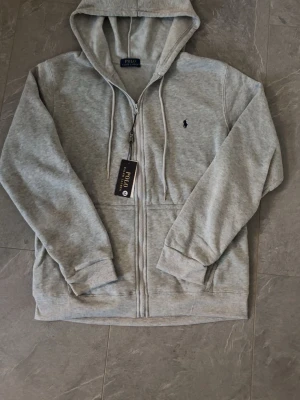 Grå hoodie från Polo Ralph Lauren - Snygg grå hoodie från Polo Ralph Lauren med dragkedja framtill, justerbar huva med snören och klassisk logga broderad på bröstet. Tillverkad i mjukt bomullsmaterial och har två fickor framtill. Perfekt för en avslappnad och stilren look.