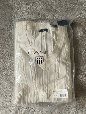 Vit stickad tröja från GANT - Kabelstickad vit tröja från GANT med V-ringning och ribbade muddar. Tröjan har en klassisk design och är tillverkad i mjukt material. Perfekt för en stilren och tidlös look. Helt ny och aldrig använd. Nypris 900kr. 