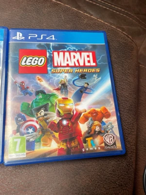 LEGO Marvel Super Heroes PS4 - LEGO Marvel Super Heroes till PS4 är ett actionäventyrsspel där du spelar som ikoniska Marvel-karaktärer i LEGO-format. Omslaget är färgglatt med Iron Man, Hulk, Spider-Man och fler hjältar. Spelet kommer i ett blått plastfodral och har PEGI 7-märkning.