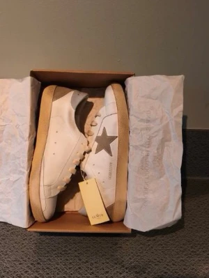 Golden Goose vita sneakers med stjärna - Säljer ett par vita Golden Goose sneakers med beige sula och grå mockastjärna på sidan. Skorna har beigea skosnören och detaljer i mocka vid hälen. Klassisk låg modell med GGDB/SSFR-tryck på sidan. Nyskick!!! 