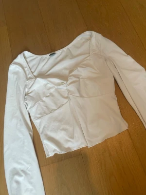 Vit långärmad croppad topp - Säljer en vit långärmad topp med croppad passform och snygga sömdetaljer över bröstet. Toppen har en modern och stilren look med en lätt stretchig känsla, perfekt att matcha med jeans eller kjol. Passar dig som gillar trendiga och enkla plagg.