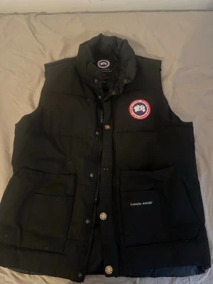 Svart dunväst från Canada Goose - Snygg svart dunväst från Canada Goose med hög krage, två stora framfickor och klassisk logotyp på bröstet. Västen har dragkedja men tyvärr så är drag grejen av  och knappar framtill men en borta  samt är vadderad för extra värme. Perfekt för lager-på-lager och streetwear-stil.