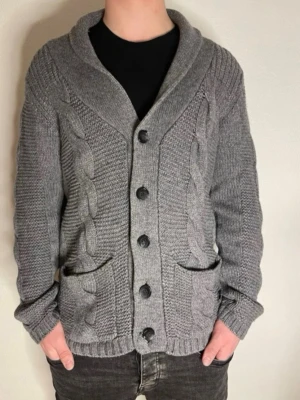 Grå stickad kofta från blueprint - Säljer en grå stickad kofta från blueprint med flätstickade detaljer framtill, stora knappar och två fickor. Koftan har sjalkrage och ribbade muddar. Perfekt för lager på lager och riktigt mysig under kyliga dagar.