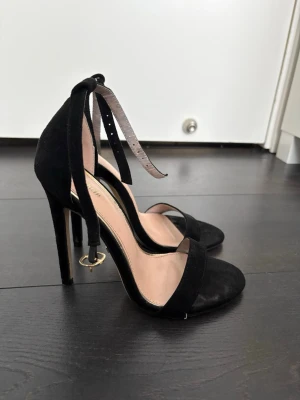 Svarta höga pumps med ankelband - Säljer ett par svarta pumps med höga klackar och snygga ankelband som knyts. Skorna har öppen tå och är tillverkade i mockaimitation med beige innersula. Perfekta för dig som vill ha en elegant och trendig look.