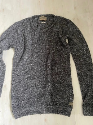 Gråmelerad stickad tröja Vaillent - Mysig gråmelerad stickad tröja från Vaillent Clothing. Tröjan har rund halsringning, lång ärm och en liten ficka framtill. Materialet är mjukt och ger en avslappnad look, perfekt för lager på lager under kyliga dagar.