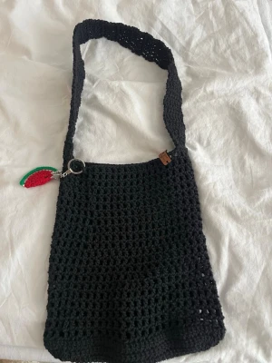 Svart virkad axelväska med charm - Säljer en svart virkad axelväska med bred axelrem och en söt nyckelring i form av en virkad vattenmelon. Väskan har en öppen, luftig struktur och en liten brun etikett på framsidan. Perfekt för dig som gillar unika accessoarer.