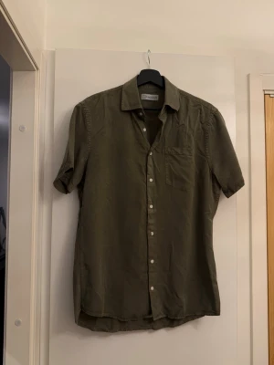 Olivgrön skjorta från Lagency - Säljer en olivgrön skjorta från Lagency i storlek S. Skjortan har korta ärmar, klassisk tröja krage, bröstficka och knappar framtill. Tillverkad i mjuk bomull som känns skön mot huden. Perfekt för en avslappnad och stilren look. I mycket bra skick, inga skador