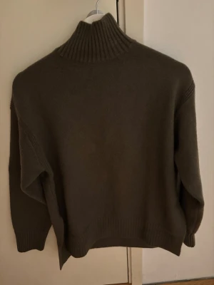 Mörkgrön  stickad polotröja - Mysig mörk grön stickad polotröja med hög ribbad krage och långa ärmar. Tröjan har en avslappnad passform och ribbade muddar vid ärmslut och nederkant. Perfekt för kyliga dagar och enkel att styla med jeans eller kjol.