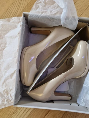 Tamaris lackpumps med hög klack - Snygga pumps i beige lack med rund tå och hög klack. Skorna har en klassisk siluett och en liten platå framtill för extra komfort. Perfekta för dig som vill ha en stilren och elegant look. Materialet är glansigt och ger en lyxig känsla. Nypris 699 kr