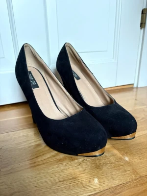 Svarta pumps med gulddetalj - Stilrena svarta pumps i mockaimitation med rund tå och hög klack. Snygg guldfärgad detalj framtill på sulan ger extra edge.