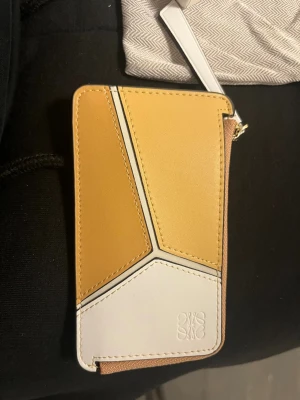 Loewe gul och vit läderplånbok - Snygg plånbok från Loewe i gult och vitt läder med geometriskt mönster. Plånboken har dragkedja, flera kortfack och präglad logga på framsidan. Perfekt för kort och kontanter, stilren och modern design. HELT NY