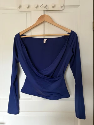 Blå omlottopp med v-ringning Nelly - Snygg blå omlottopp med djup v-ringning och långa ärmar. Toppen har en figurnära passform och är tillverkad i ett mjukt, stretchigt material som känns bekvämt mot huden.