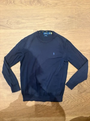 Mörkblå stickad tröja Polo Ralph Lauren - Mörkblå stickad tröja från Polo Ralph Lauren i slim fit-modell. Tillverkad av mjuk pima cotton och har rund halsringning samt den klassiska broderade loggan på bröstet. Perfekt för dig som gillar stilrena och tidlösa plagg. Den är i storlek S.