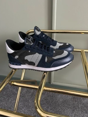 Valentino Garavani camo sneakers - Snygga sneakers från Valentino Garavani med mörkblått och svart camo-mönster, vita detaljer och meshpaneler. Skorna har klassisk låg siluett, svarta snören och robust sula. Perfekta för dig som vill sticka ut med stilrena detaljer och exklusiv känsla. Äkta fick i present men ville inte ha dem mer så säljer dem! Är storlek 43 men sitter som 44 typ.