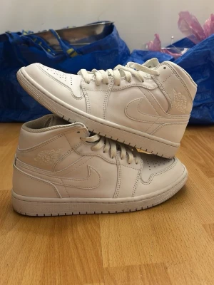 Vita Air Jordan 1 Mid sneakers - Säljer ett par fräscha vita Air Jordan 1 Mid sneakers från Nike. Skorna har klassisk siluett, perforerad tåbox och ikoniska vingar-loggan på sidan. Tillverkade i slätt läder med matchande vita skosnören och sula. Perfekta för dig som gillar stilrena och tidlösa sneakers. Kan diskutera pris