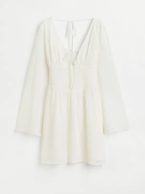 Vit kortklänning med knyt och v-ringning - Supersöt vit kortklänning med v-ringning och knytband både fram och bak. Klänningen har långa, lätt transparenta ärmar och ett flowy fall. Perfekt för sommarens alla tillfällen och ger en romantisk vibe. Tillverkad i ett tunt, mjukt material som känns luftigt.