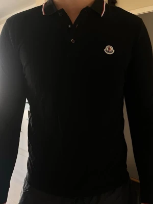 Moncler piké - Snygg svart långärmad piké från Moncler med klassisk krage och knappslå. Diskret Moncler-logga på bröstet och tunna röda och vita detaljer på kragen. Perfekt för en clean och stilren look.