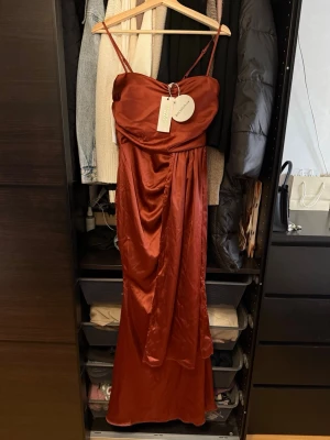 Röd långklänning från Boohoo - Snygg röd långklänning från Boohoo med tunna axelband och draperad detalj framtill. Klänningen har en elegant, glansig finish och en figurnära passform som ger en lyxig känsla. Perfekt för dig som vill sticka ut med en stilren look.