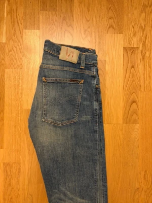 Blå slim fit jeans från Nudie Jeans - Säljer ett par klassiska blå slim fit jeans från Nudie Jeans med snygg slitning och kontrastsömmar. Jeansen har fem fickor, normal midja och är tillverkade i mjuk denim. Perfekta för en avslappnad och trendig look.