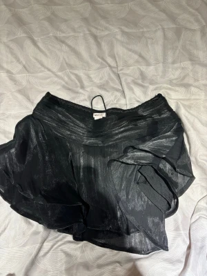 Svart omlottkjol med volanger - Säljer en svart kort omlottkjol från Gina Tricot med volangdetaljer. Kjolen har ett glansigt tyg och elastisk midja som ger en bekväm passform. Perfekt för dig som vill ha en trendig och snygg kjol till sommaren.