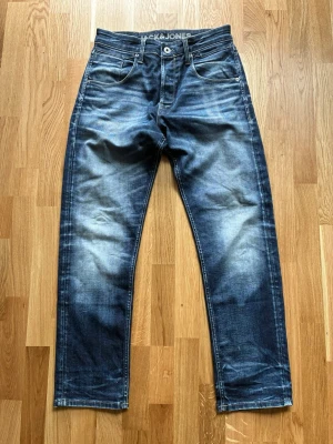Blå raka jeans från Jack & Jones - Säljer ett par blå denimjeans från Jack & Jones med snyggt slitna detaljer och raka ben. Klassisk femficksmodell med knappgylf och tydliga kontrastsömmar. Perfekta för en avslappnad stil.
