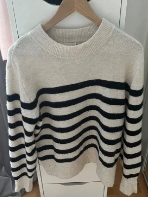Beige stickad tröja med svarta ränder - Stickad tröja i beige med breda svarta ränder på ärmarna. Rund halsringning och ribbstickade muddar. Perfekt för dig som gillar en enkel men trendig look. Materialet är mjukt och bekvämt.