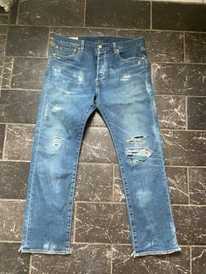 Blå slitna Levi's jeans med hål - Säljer ett par blå Levi's jeans med raka ben och slitningar samt hål på framsidan för en cool, avslappnad look. Klassisk femficksmodell med läderpatch bak i midjan och råa benslut. Perfekta för dig som gillar streetstyle och vintage vibe.