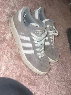 Grå Adidas Campus sneakers - Säljer ett par grå Adidas Campus sneakers med klassiska vita ränder på sidorna och vit sula. Skorna har lågt skaft och är gjorda i mocka med textildetaljer. Perfekta för dig som gillar en clean och tidlös stil.
