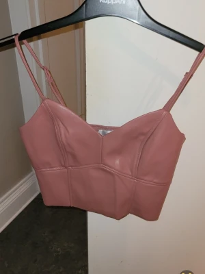 Rosa linne i skinnimmitation med smala band stl S - ANV 1 G Snygg rosa linnetopp i skinnimitation med smala axelband och v-ringad hals. STL S Toppen är croppad och har en stilren design som passar perfekt till höga jeans eller kjol. Perfekt för dig som vill ha en trendig och edgy look reglerbara band , borstad insida Längd från axeln och ner ca 39 cm Längd linne utan axelband 27 cm Mått över byst 34 cm ( 68 cm runtom ) Dragkedja i sidan , från Newyorker 