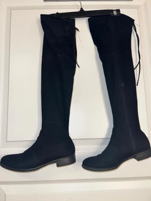 Svarta overknee stövlar i mocka - Snygga svarta overknee stövlar i mjuk mocka med låg klack och rund tå. Stövlarna har dragkedja på insidan och snörning upptill för perfekt passform. Perfekta till höstens och vinterns outfits.