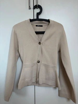 Beige kofta från Gina Tricot - Snygg beige kofta från Gina Tricot med två stora knappar framtill och två fickor. Koftan har v-ringning, långa ärmar och ribbade muddar. Tillverkad i mjukt stickat material som känns skönt mot huden. Perfekt för lager på lager och enkel att matcha.