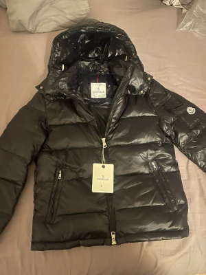 Svart dunjacka från Moncler med huva - Säljer en svart dunjacka från Moncler med avtagbar huva, två dragkedjefickor och klassisk Moncler-logga på ärmen. Jackan har en glansig finish och breda kanaler som ger en modern look. Perfekt för kalla vinterdagar.