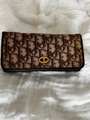 Vintage Brun Dior monogram handväska - Vintage Dior -monogramväska i bruna nyanser med läderdetaljer. Guldfärgad CD-logotyp framstill. Klaff bäras över axeln eller axelband en guldfärgad kedja (extra, omärkt) Användningsspår: några märken på fodret, logotypen på insidan har blekts. Mått: bredd 18 cm, höjd 9 cm, djup 3 cm. Kedjans längd 100cm. Perfekt för dig som gillar lyxiga accessoarer och tidlös design. Kontakta mig endast vid seriösa intressen och för fler bilder. 