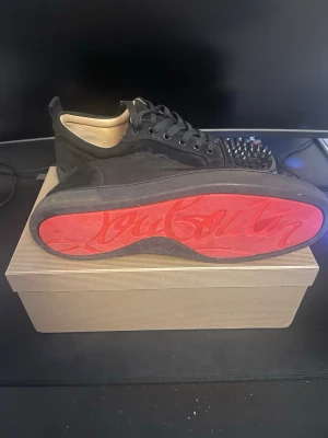 ÄKTA Christian Louboutin svarta sneakers med nitar - Säljer ett par svarta sneakers från Christian Louboutin i mocka med ikonisk röd sula och svarta nitar på tån. Skorna har snörning och rund tå, samt beige innersula. Grymt statement-plagg för dig som gillar edgy detaljer. Priset på grund av skicket av skorna 