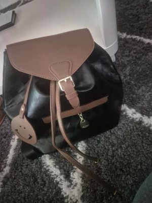 Svart och brun ryggsäck med spänne - Snygg ryggsäck i svart med bruna detaljer och lock. Väskan har ett justerbart spänne framtill, dragkedjeficka och dekorativt hängsmycke. Materialet ser ut att vara konstläder och textil. Perfekt för dig som vill ha en stilren och praktisk väska.