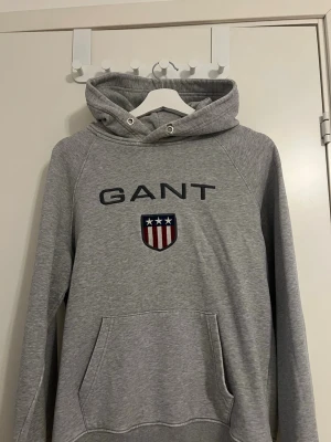 Grå hoodie från GANT med logga - Snygg grå hoodie från GANT med broderad logga och sköld med stjärnor och ränder på bröstet. Klassisk känguruficka framtill och justerbar huva med dragsko. Tillverkad i mjukt bomullsmaterial som är skönt att ha på sig. De defekter som finns på plagget är att snörena är borta, annars felfritt! 